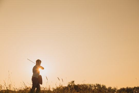 Black silhouette of cute young caucasian kid isolated on sunny orange sunset or sunrise sky background playing sword with invisible enemy. Horizontal color photography.