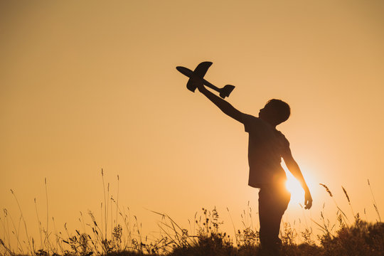 Black silhouette of young caucasian kid isolated on sunny golden sunset sky background. Boy playing toy plane outside on grassy summer hill. Setting big goals and dreaming about happy future concept.