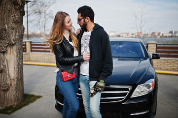 Fototapeta premium Cool multiracial couple hugs together against black elegant sport car.