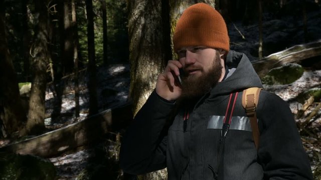 A bearded man in a hat talking on a cell phone in a coniferous forest. The concept of cellular communication in hard to reach places