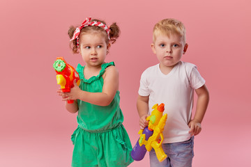 Happy little toddler boy and girl  in summer clothes holds toy water guns isolated on pink wall background. Children studio portrait. People childhood lifestyle concept. 
