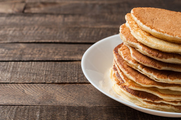 Heap of homemade pancakes without cream lies on round white ceramic plate and old rustic wooden table. Copy space for text