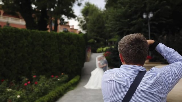 professional photographer in striped shirt makes photos of wedding couple in green garden slow motion backside view