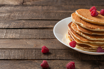 Heap of homemade pancakes with fresh raspberry lies on round white ceramic plate and old rustic wooden table. Copy space for text