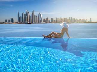 Beautiful panorama of Dubai Marina skyline in a background with a pool, deck chair and woman with a white hat.
