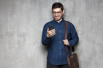 Modern smiling businessman in glasses and denim shirt standing against gray textured wall with smartphone in hand and bag on shoulder