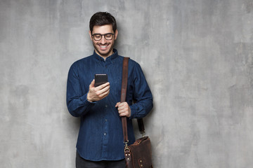 Modern smiling entrepreneur in glasses and denim shirt standing against gray textured wall with...