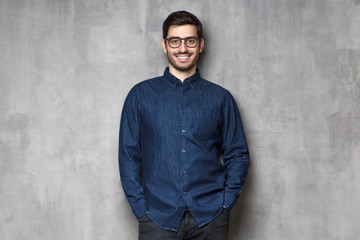 Young handsome man wering eyeglasses, smiling and feeling confident, standing against gray textured wall background