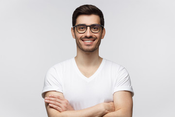 Fototapeta premium Portrait of smiling young man in casual white t-shirt, looking at camera, standing with crossed arms isolated on gray background