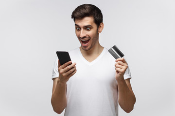 Young happy man holding smartphone and credit card in hands, looking at screen with amazement,...