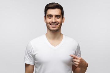 Studio shot of smiling young man pointing at his blank white t-shirt with index finger, copy space for your ads, isolated on gray background