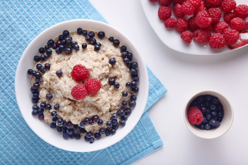 Flat top view of homemade vegan breackfast, picture of plate with oatmeal and fresh fruit berries on white kitchen table, breakfast on blue towel on desk. Healthy eating and health care concept.
