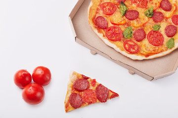Photo of pepperoni pizza with mozzarella cheese, salami, pepper, spices and fresh spinach, cardboard box, piece of pizza and three tomatoes isolated on white background. Fast food eating concept.