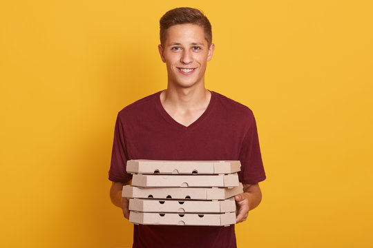 Image of joyful handsome delivery man wearing casual burgundy t shirt, holding stack of pizza boxes in hands and looking directly at camera isolated over yellow studio background. Junk food concept.