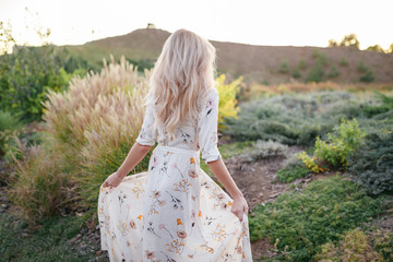Back view blound young woman wearing long dress walking in the park. Beautiful young girl walking...