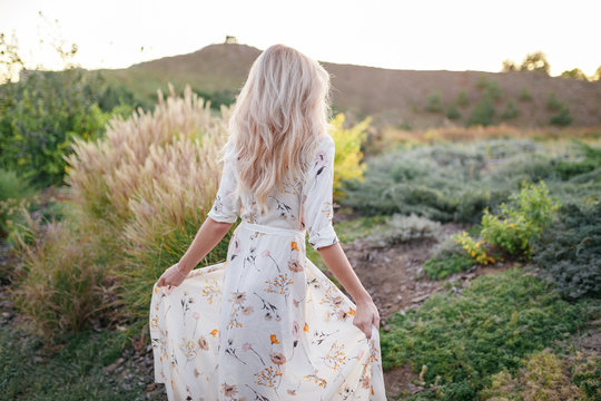 Back view blound young woman wearing long dress walking in the park. Beautiful young girl walking in a field