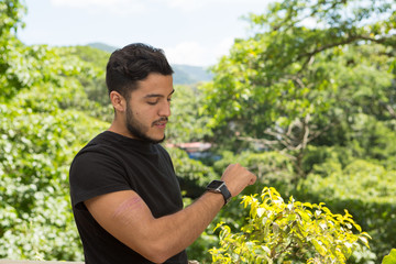 Naklejka na ściany i meble Young latin man portrait indoors next to a window with nature as background. He is smiling He is using the smartwatch. his is wearing.