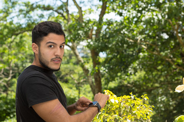 Naklejka na ściany i meble Young latin man portrait indoors next to a window with nature as background. He is smiling He is using the smartwatch. his is wearing.