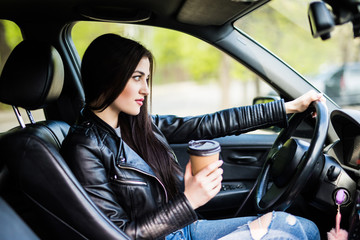 Fototapeta na wymiar Beautiful girl drinking coffee, looking at camera and smiling while sitting in her car