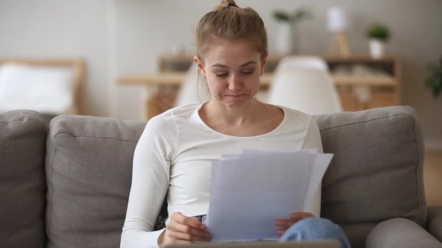Young woman siting on couch holding documents papers feels frustrated