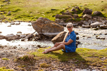 happy beautiful retirement age woman in blue dress sitting on the grass near the river