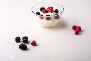 Semolina pudding porridge dessert with  blackberry and raspberry in glass bowl near with scattered heap berries on white wooden background, breakfast, isolated