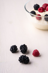 Semolina pudding porridge dessert with  blackberry and raspberry in glass bowl near with scattered heap berries on white wooden background, breakfast, isolated