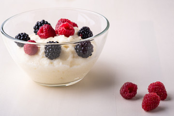 Semolina pudding porridge dessert with  blackberry and raspberry in glass bowl near with scattered heap berries on white wooden background, breakfast, isolated