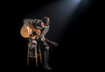 musician playing acoustic guitar, sitting on high chair, black background with beautiful soft light
