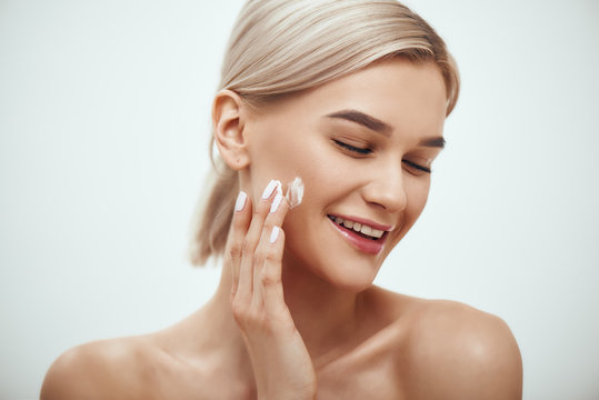 Portrait of cute blonde woman spreading cream on her face and smiling while standing against grey background. Skin care