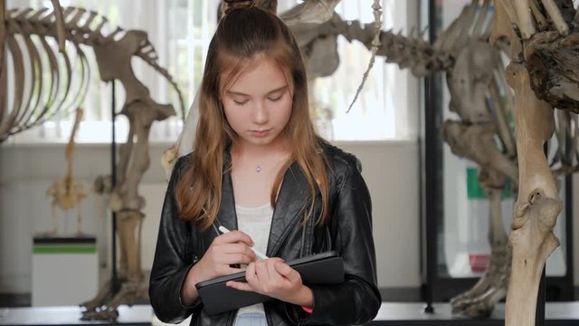 Teenager cute girl makes draw sketches in graphic tablet of animal skeletons in museum of nature as part of school task of biology