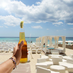 Male hand holding a bottle of beer against a sunny sky and crystal clear sea. In the background a beautiful beach resort. Vacation concept.