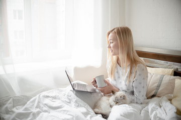 Obraz premium Work from home concept . Blonde Giirl typing and using laptop. Hygge with fluffy cat, pajama and coffee cup. Horizontal copyspace