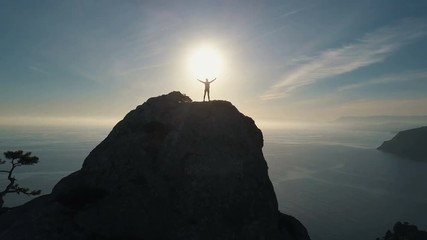 Aerial silhouette of a young woman climbing to the top of a mountain against the sea at sunset. Lady on the summit in beautiful scenery raises her hands up.
