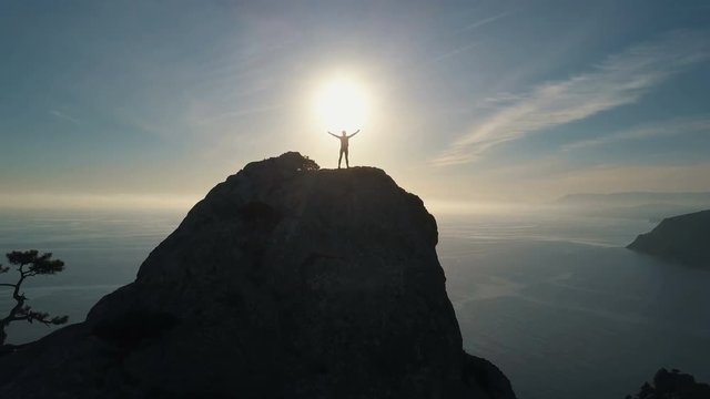 Aerial silhouette of a young woman climbing to the top of a mountain against the sea at sunset. Lady on the summit in beautiful scenery raises her hands up.