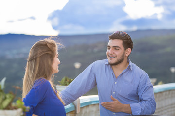 Couple having nice conversation outside. They are latin in their twenties. They both wear casual clothes. The focus is on him who is in front of the camera.