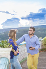 Couple having nice conversation outside. They are latin in their twenties. They both wear casual clothes. The focus is on him who is in front of the camera.