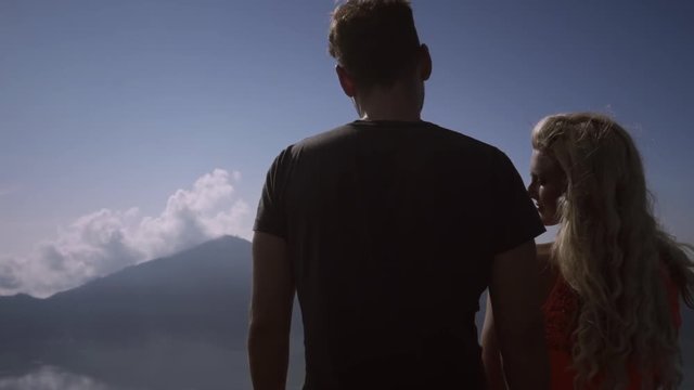 Young woman in pink dress with man looking on amazing view from top of volcano. Beautiful couple standing at edge turns head on background of mountains, valley, lake and sky.