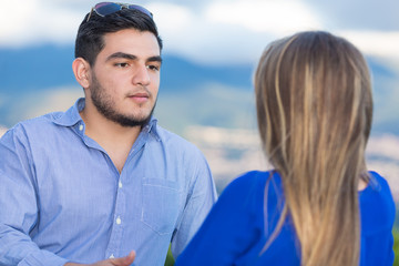 Man having conversation with woman outside. They are latin in their twenties. They both wear casual clothes. The focus is on him who is in front of the camera.