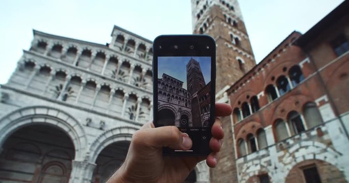 Close-up man hand holding smartphone taking pictures on camera. Male tourist takes a photo of famous Torre Guinigi stunning landmark in center of Lucca city. Tour to Italy.