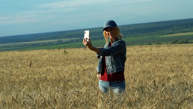 A slender woman walks on a wheat field and takes pictures of spikelets on her phone on a Sunny summer day.
