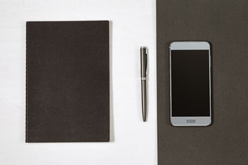 Black cover of closed notebook and silver pen. Top view on smartphone above office desk background. Working space. Flat lay.