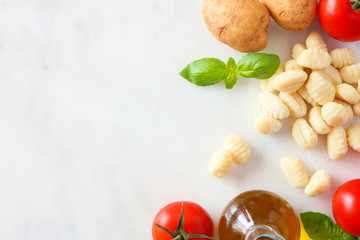 Gnocchi meal ingredients including fresh gnocchi, tomatoes, olive oil, potatoes and basil. Top view, side border against a white marble background. Copy space.