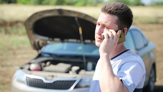 A young, handsome сaucasian driver stands next to a broken car and calls the service center, close-up