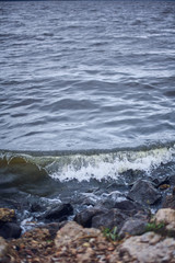 Fototapeta na wymiar Small wave on the stone shore. Sea or lake with dark water. Wild Nature Dramatic Background