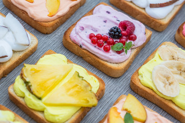 Assorted toasts with fruits and berries on wipped cream on a wood board on light background. Summer traditional homemade dessert.