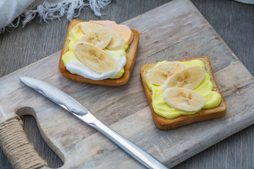Toast with banana on wipped cream on a wood board on light background. Summer traditional homemade dessert.