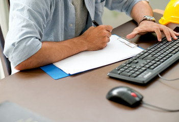 Engineer man is writing something on the paper while typing on keyboard of computer with concept of working at home.