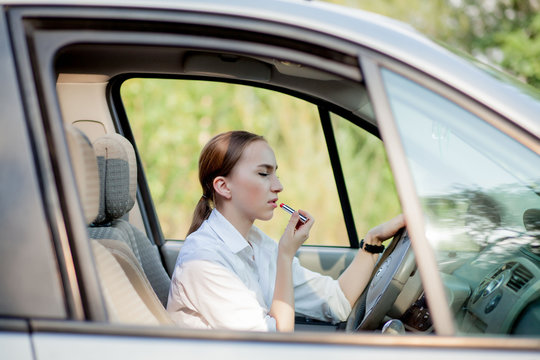 Concept of danger driving. Young woman driver red haired teenage girl painting her lips doing applying make up while driving the car