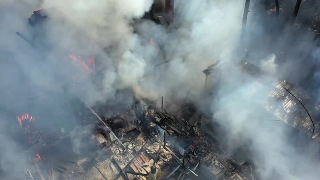 Firefighters pour water from fire hoses on the burning roof, smoke, protruding chimney. Disaster fire. Summer day. Close-up from the drone .